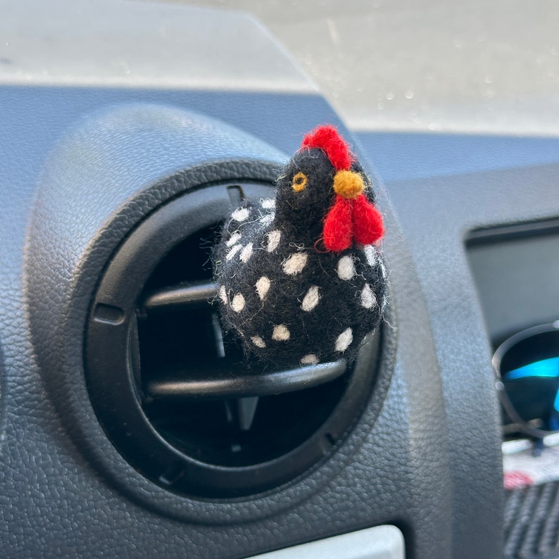 Puede incluir: Un ambientador con forma de gallina negra con lunares blancos, cresta roja y pico amarillo, enganchado a una rejilla de ventilaci&oacute;n de un coche. La gallina est&aacute; hecha de un material suave y a&ntilde;ade un toque decorativo al interior del veh&iacute;culo.