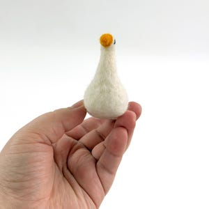 K&ouml;nnte beinhalten: Eine kleine, cremewei&szlig;e, nadelgefilzte G&auml;nsefigur mit rundlichem K&ouml;rper, schlankem Hals und einem leuchtend orangefarbenen Schnabel. Die Textur der Wolle ist auf der gesamten Oberfl&auml;che des Vogels sichtbar, der eine einfache Form aufweist.