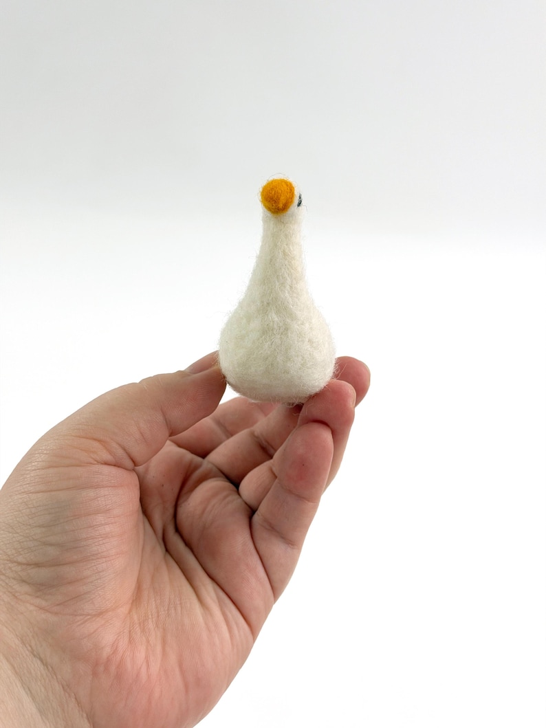K&ouml;nnte beinhalten: Eine kleine, cremewei&szlig;e, nadelgefilzte G&auml;nsefigur mit rundlichem K&ouml;rper, schlankem Hals und einem leuchtend orangefarbenen Schnabel. Die Textur der Wolle ist auf der gesamten Oberfl&auml;che des Vogels sichtbar, der eine einfache Form aufweist.