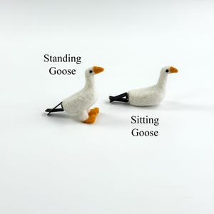 K&ouml;nnte beinhalten: Zwei wei&szlig;e Haarspangen in G&auml;nseform aus Filz. Eine ist mit Standing Goose beschriftet und zeigt eine stehende Gans mit orangen F&uuml;&szlig;en. Die andere ist mit Sitting Goose beschriftet und zeigt eine sitzende Gans. Beide haben schwarze Augen, orange Schn&auml;bel und schwarze Metallclips.
