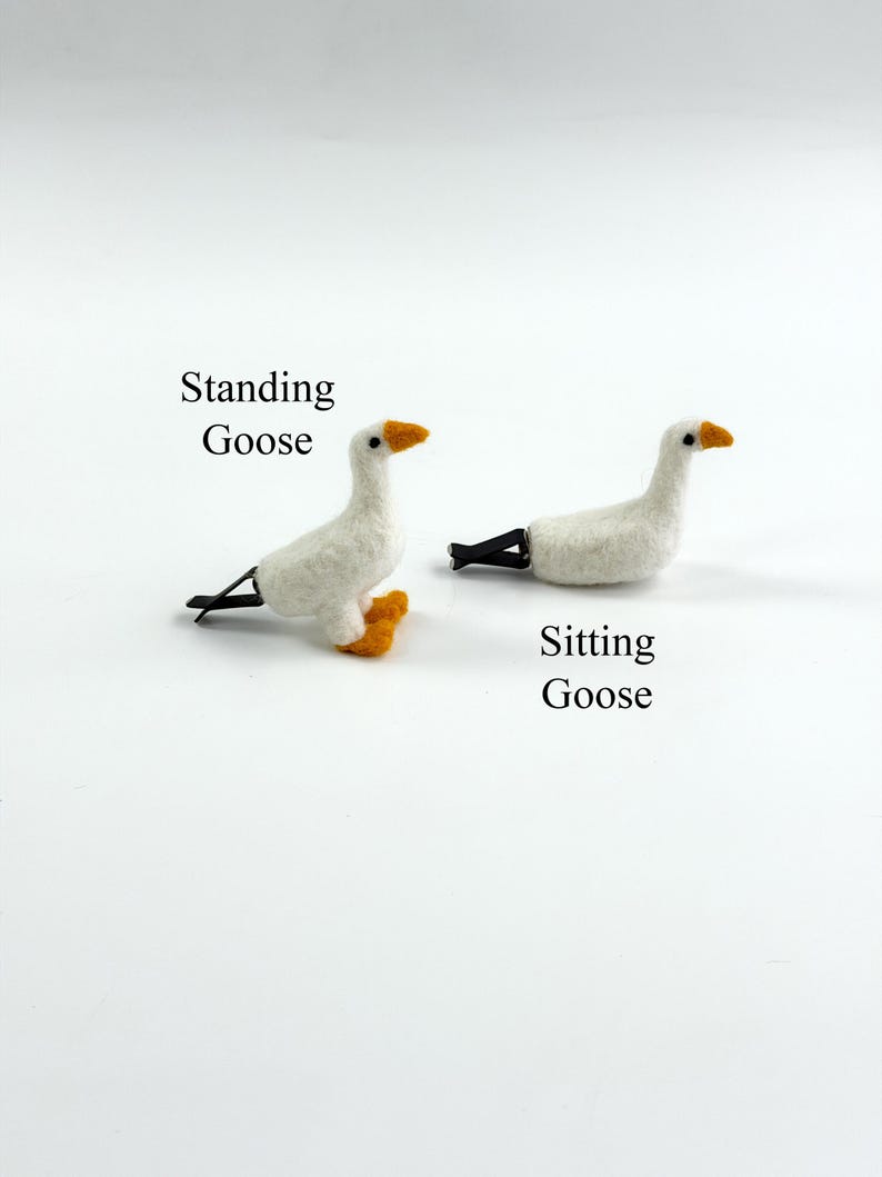 K&ouml;nnte beinhalten: Zwei wei&szlig;e Haarspangen in G&auml;nseform aus Filz. Eine ist mit Standing Goose beschriftet und zeigt eine stehende Gans mit orangen F&uuml;&szlig;en. Die andere ist mit Sitting Goose beschriftet und zeigt eine sitzende Gans. Beide haben schwarze Augen, orange Schn&auml;bel und schwarze Metallclips.
