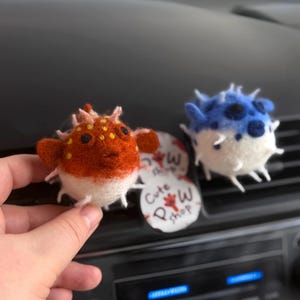 Op de afbeelding: Twee handgemaakte kogelvisornamenten van vilt. Eén is oranje en wit met zwarte stippen, de andere is blauw en wit. Een kleine ronde sticker met de tekst "Cute P Shop" is ook zichtbaar.