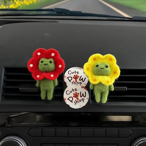 May include: Two felt frog figurines with flower-shaped heads, one red with white spots and the other yellow. They are standing on a car dashboard next to two round stickers with the text "Cute Paw Shop". The background shows a road.