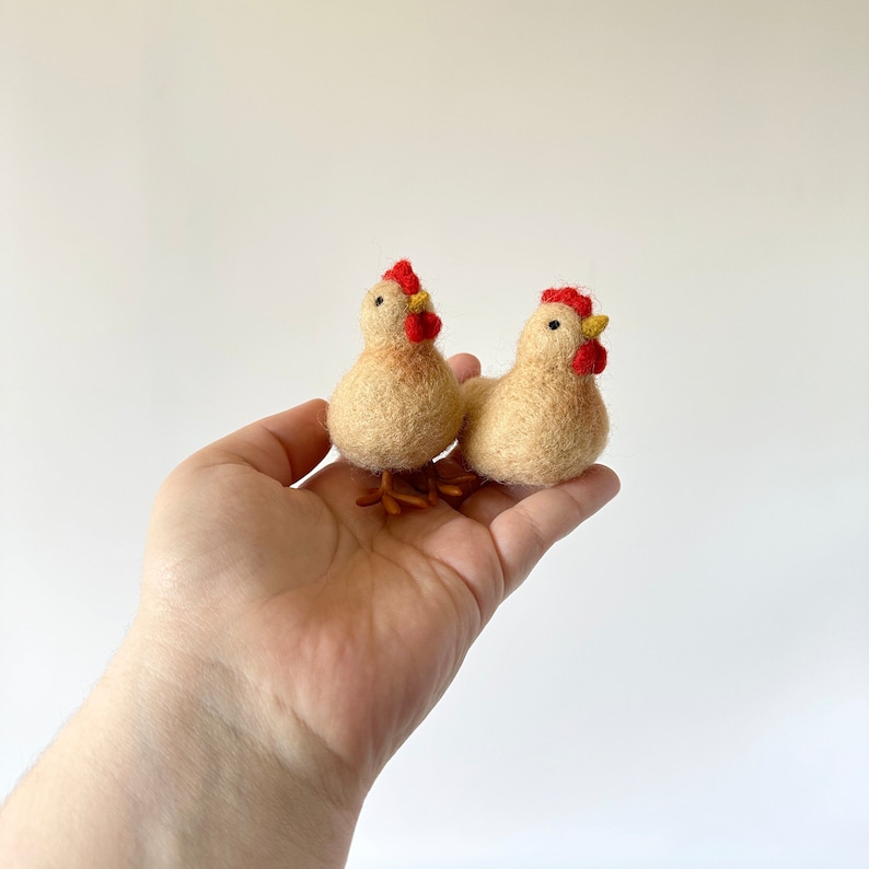 May include: Two small felt chickens, one beige and one brown, with red combs and yellow beaks. The chickens are sitting in a hand.