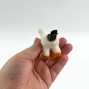 K&ouml;nnte beinhalten: Eine kleine, wei&szlig; nadelgefilzte G&auml;nsefigur mit leuchtend orangefarbenen F&uuml;&szlig;en. Auf dem R&uuml;cken der Gans ist eine schwarze Metall-Krokodilklemme befestigt, wodurch sie als dekorative Klammer oder Zubeh&ouml;r zum Halten von Papieren verwendet werden kann.