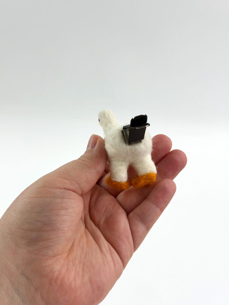 K&ouml;nnte beinhalten: Eine kleine, wei&szlig; nadelgefilzte G&auml;nsefigur mit leuchtend orangefarbenen F&uuml;&szlig;en. Auf dem R&uuml;cken der Gans ist eine schwarze Metall-Krokodilklemme befestigt, wodurch sie als dekorative Klammer oder Zubeh&ouml;r zum Halten von Papieren verwendet werden kann.