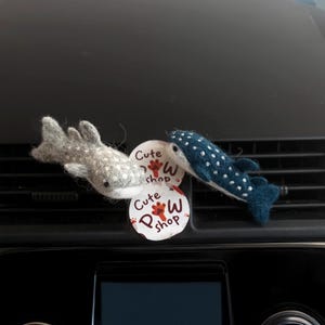 Könnte beinhalten: Zwei Wal-Ornamente aus Filz, eines grau mit weißen Punkten und eines blau mit weißen Punkten, liegen auf dem Armaturenbrett eines Autos. Ein runder Aufkleber mit dem Text "Cute Paw Shop" ist ebenfalls sichtbar.