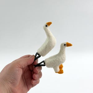 K&ouml;nnte beinhalten: Zwei nadelgefilzte wei&szlig;e G&auml;nsefiguren mit orangefarbenen Schn&auml;beln und F&uuml;&szlig;en, die jeweils an einer schwarzen Metallklammer befestigt sind. Die G&auml;nse haben kleine schwarze Perlenaugen und eine weiche, texturierte Wolloptik.