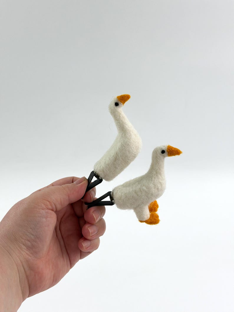 K&ouml;nnte beinhalten: Zwei nadelgefilzte wei&szlig;e G&auml;nsefiguren mit orangefarbenen Schn&auml;beln und F&uuml;&szlig;en, die jeweils an einer schwarzen Metallklammer befestigt sind. Die G&auml;nse haben kleine schwarze Perlenaugen und eine weiche, texturierte Wolloptik.