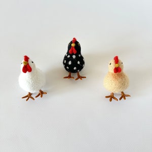 Puede incluir: Tres figuras de gallina de fieltro. Una es blanca, otra negra con lunares blancos y la tercera amarillo claro. Cada una tiene una cresta y una barbilla rojas, y patas marrones. Estas gallinas decorativas est&aacute;n sobre un fondo blanco.