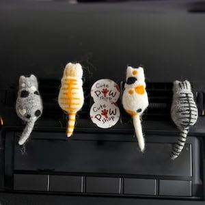 May include: Four handmade felt cat air fresheners in a car. The cats are white, grey, and orange with black and grey stripes and spots. Two circular white stickers with the text "Cute Paw Shop" are also visible.