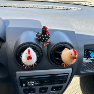 May include: Three felt chicken figurines, one black and white, one white, and one peach, are displayed in a car's air vent.