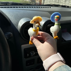 May include: Two felt goose figurines, one with a yellow hat and scarf, the other with a blue hat and green scarf. The geese have orange feet and beaks. One goose holds a small flower. The figurines are white with black eyes.