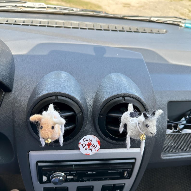 以下が含まれることがあります： 車のエアコン吹き出し口にぶら下がっている、動物の形をした2つの毛糸の車の芳香剤。動物は白と茶色の羊と黒と白の牛。羊は左側に、牛は右側にあります。羊の下には「Cute Paw Shop」と書かれた赤と白のステッカーがあります。