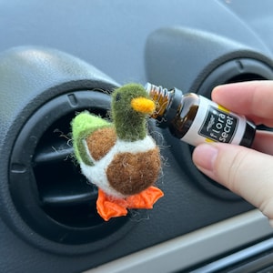Könnte beinhalten: Ein Filz-Enten-Lufterfrischer für das Auto mit orangenen Füßen und einem grün-braun-weißen Muster. Eine Hand hält eine kleine braune Glasflasche mit einem gelben Etikett, auf dem "flora secret" steht, und gießt ätherisches Öl in den Lufterfrischer.
