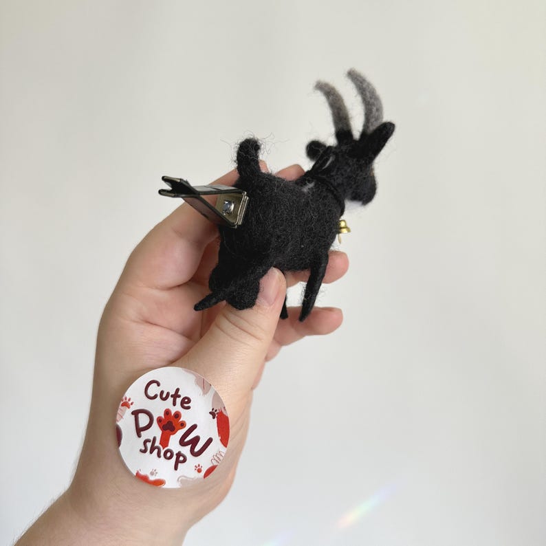以下が含まれることがあります： 黒いフェルトのヤギで、背中にクリップが付いています。ヤギは首にベルが付いていて、手に乗っています。手は「Cute Paw Shop」と書かれた白いステッカーを持っています。