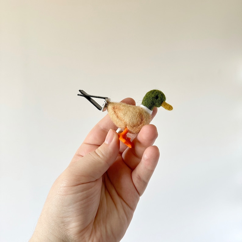 K&ouml;nnte beinhalten: Ein kleiner Filz-Enten-Anh&auml;nger mit gr&uuml;nem Kopf, wei&szlig;em Kragen und orangefarbenen F&uuml;&szlig;en. Die Ente ist an einer schwarzen Metallklammer befestigt.