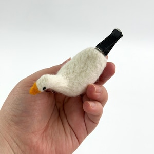 K&ouml;nnte beinhalten: Eine nadelgefilzte wei&szlig;e G&auml;nsefigur mit einem leuchtend orangefarbenen Schnabel und einem kleinen schwarzen Auge. Die Gans ist an einem schwarzen Kunststoffclip befestigt, der dazu dient, das Objekt zu Dekorationszwecken an einer L&uuml;ftung zu befestigen.