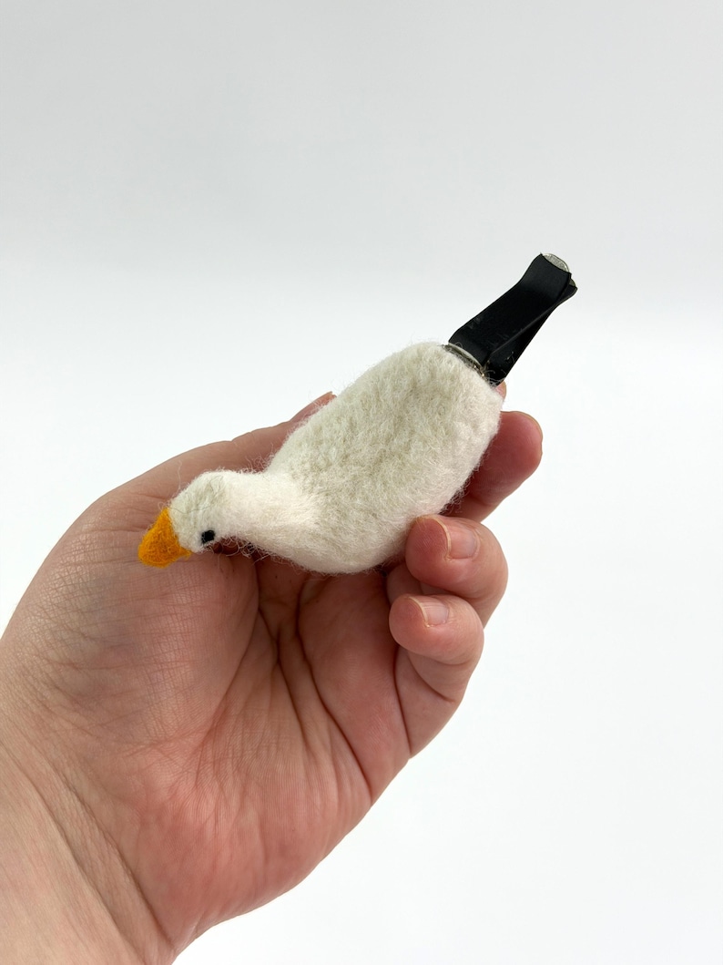 K&ouml;nnte beinhalten: Eine nadelgefilzte wei&szlig;e G&auml;nsefigur mit einem leuchtend orangefarbenen Schnabel und einem kleinen schwarzen Auge. Die Gans ist an einem schwarzen Kunststoffclip befestigt, der dazu dient, das Objekt zu Dekorationszwecken an einer L&uuml;ftung zu befestigen.