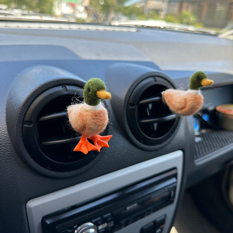 K&ouml;nnte beinhalten: Zwei Filz-Enten-Lufterfrischer, die an den L&uuml;ftungsschlitzen des Armaturenbretts eines Autos h&auml;ngen. Die Enten sind braun und wei&szlig; mit gr&uuml;nen K&ouml;pfen und orangefarbenen F&uuml;&szlig;en.