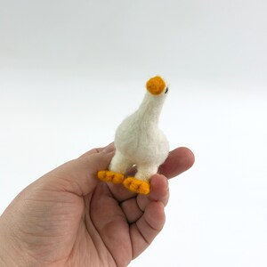 K&ouml;nnte beinhalten: Eine kleine, handgefertigte, nadelgefilzte G&auml;nsefigur aus wei&szlig;er Wolle. Die Gans hat einen runden K&ouml;rper, einen langen Hals und einen kleinen Kopf mit einem leuchtend orangefarbenen Schnabel. Sie steht auf zwei kleinen, flachen, orangefarbenen F&uuml;&szlig;en.