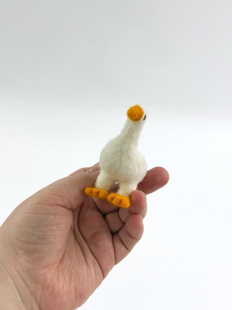 K&ouml;nnte beinhalten: Eine kleine, handgefertigte, nadelgefilzte G&auml;nsefigur aus wei&szlig;er Wolle. Die Gans hat einen runden K&ouml;rper, einen langen Hals und einen kleinen Kopf mit einem leuchtend orangefarbenen Schnabel. Sie steht auf zwei kleinen, flachen, orangefarbenen F&uuml;&szlig;en.