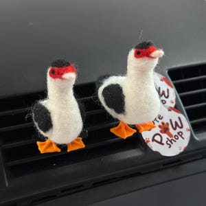 Op de afbeelding: Twee handgemaakte vilten eendenornamenten. De eenden hebben witte lichamen, zwarte vlekken, rode petten en oranje voeten. De ene eend is iets kleiner dan de andere. De eenden worden tentoongesteld op een ventilatierooster van een auto. Een label met de tekst "P W shop" is zichtbaar.