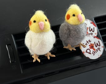 Può includere: Due ornamenti a forma di uccello in feltro, uno bianco e giallo, l'altro grigio e giallo, con macchie arancioni sulle guance e becchi rosa. Gli uccelli sono appollaiati su una presa d'aria dell'auto. È visibile un'etichetta circolare con la scritta "Cute Paw Shop".