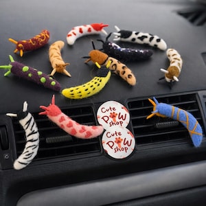 Könnte beinhalten: Eine Sammlung von filzartigen, schneckenförmigen Auto-Lufterfrischern. Die Schnecken weisen verschiedene Muster und Farben auf, darunter Streifen, Punkte und Herzen. Zwei runde Aufkleber mit dem Text "Cute Paw Shop" sind ebenfalls zu sehen.