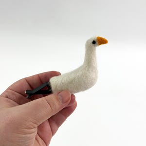 K&ouml;nnte beinhalten: Eine nadelgefilzte wei&szlig;e G&auml;nsefigur mit strukturiertem Wollk&ouml;rper, einem kleinen orangefarbenen Schnabel und einem schwarzen Auge. Die Gans ist an einem schwarzen Kunststoffclip befestigt, der f&uuml;r die Anbringung an einer L&uuml;ftung gedacht ist.
