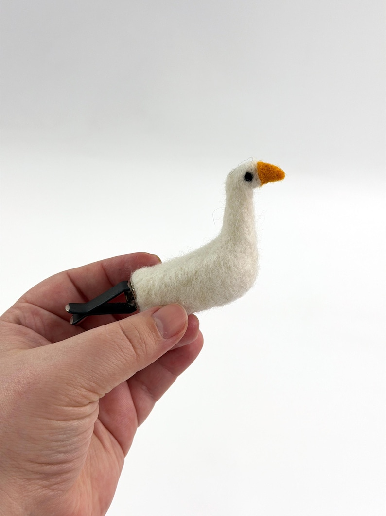 K&ouml;nnte beinhalten: Eine nadelgefilzte wei&szlig;e G&auml;nsefigur mit strukturiertem Wollk&ouml;rper, einem kleinen orangefarbenen Schnabel und einem schwarzen Auge. Die Gans ist an einem schwarzen Kunststoffclip befestigt, der f&uuml;r die Anbringung an einer L&uuml;ftung gedacht ist.