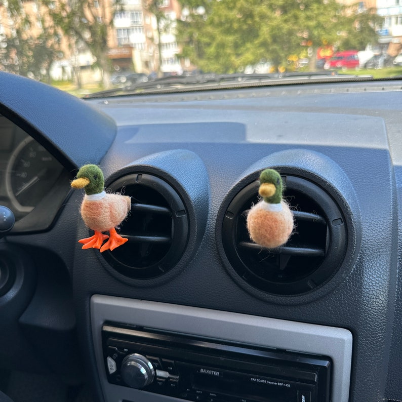 K&ouml;nnte beinhalten: Zwei Filz-Enten-Lufterfrischer, die an den L&uuml;ftungsschlitzen eines Autos h&auml;ngen. Die Enten sind braun und gr&uuml;n mit orangenen F&uuml;&szlig;en.