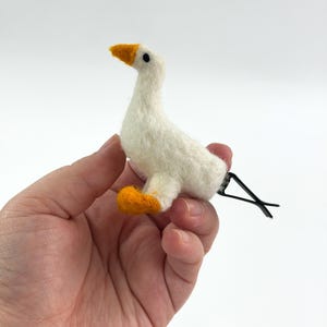 K&ouml;nnte beinhalten: Eine nadelgefilzte wei&szlig;e G&auml;nse-Haarspange mit einem strukturierten cremewei&szlig;en K&ouml;rper, einem leuchtend orangefarbenen Schnabel und orangefarbenen F&uuml;&szlig;en. Die kleine G&auml;nsefigur ist an einer schwarzen Metall-Krokodilklemme zur sicheren Befestigung befestigt.