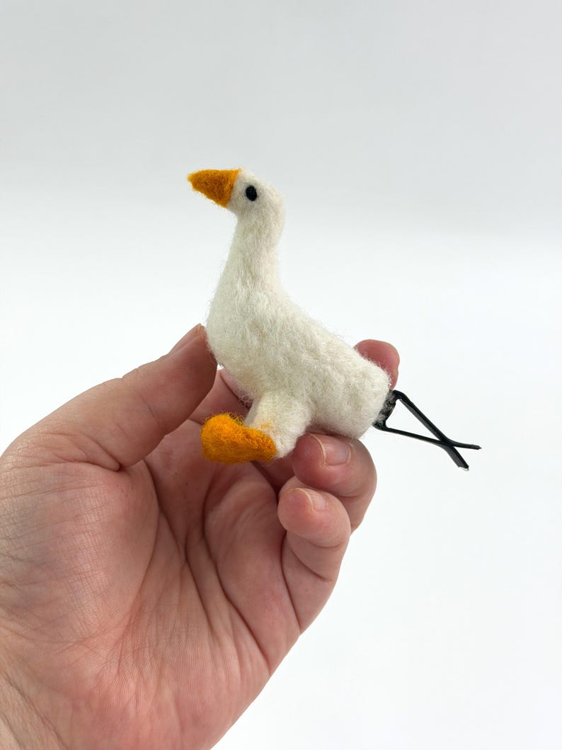 K&ouml;nnte beinhalten: Eine nadelgefilzte wei&szlig;e G&auml;nse-Haarspange mit einem strukturierten cremewei&szlig;en K&ouml;rper, einem leuchtend orangefarbenen Schnabel und orangefarbenen F&uuml;&szlig;en. Die kleine G&auml;nsefigur ist an einer schwarzen Metall-Krokodilklemme zur sicheren Befestigung befestigt.