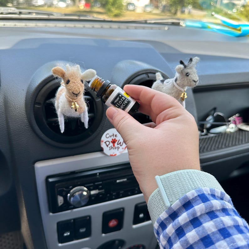 以下が含まれることがあります： 車のエアコン吹き出し口にぶら下がっている、茶色と白のニット製の小さなヤギの置物。手は、白いラベルに「floral secret」と書かれた小さな透明なガラス瓶を持っている。