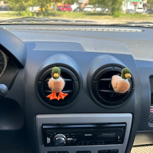 K&ouml;nnte beinhalten: Zwei Filz-Enten-Lufterfrischer, die an den L&uuml;ftungsschlitzen des Armaturenbretts eines Autos h&auml;ngen. Die Enten sind braun mit orangenen F&uuml;&szlig;en und gr&uuml;nen K&ouml;pfen.