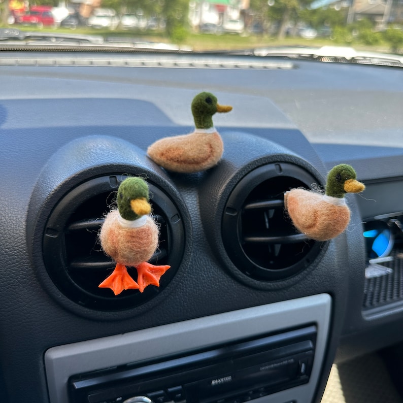 K&ouml;nnte beinhalten: Drei Filz-Enten-Lufterfrischer f&uuml;r das Auto. Die Enten sind braun mit gr&uuml;nen K&ouml;pfen und orangefarbenen F&uuml;&szlig;en. Sie befinden sich in den L&uuml;ftungsschlitzen eines Armaturenbretts.
