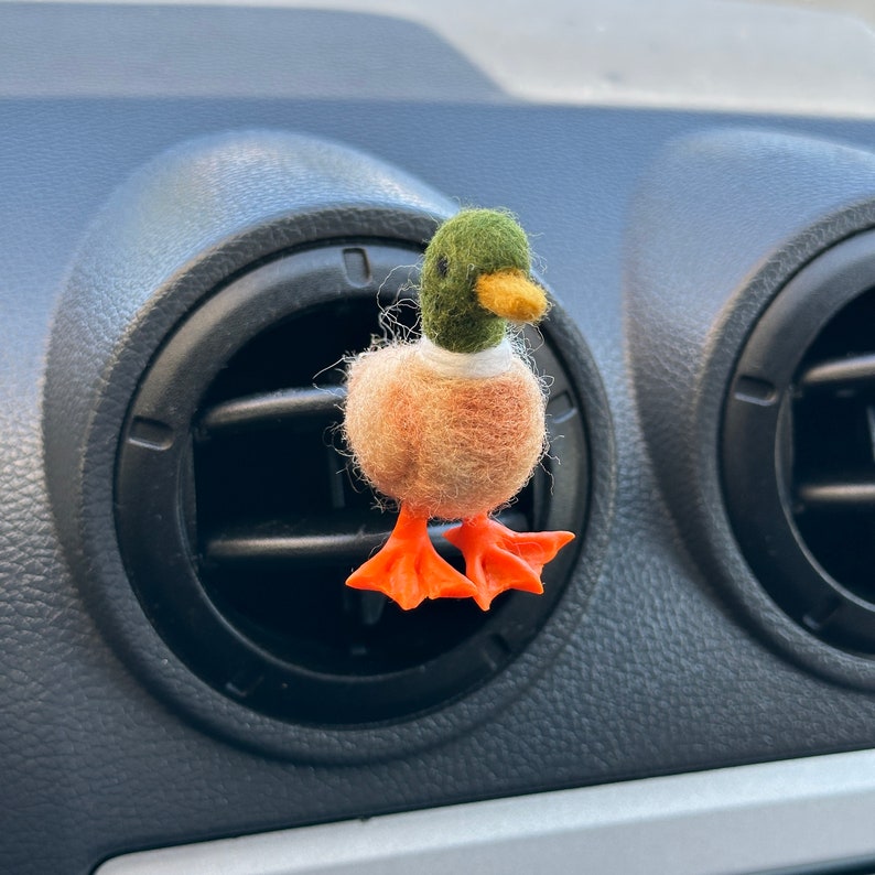 K&ouml;nnte beinhalten: Ein Filz-Enten-Lufterfrischer f&uuml;r das Auto mit gr&uuml;nem Kopf, braunem K&ouml;rper und orangefarbenen F&uuml;&szlig;en. Die Ente h&auml;ngt an einer L&uuml;ftung im Auto.