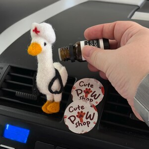 May include: A felted white goose wearing a nurse's cap with a red cross, stethoscope, and orange beak and feet. Two round stickers with the text "Cute Paw Shop" and a paw print are visible. A small brown bottle is held nearby.