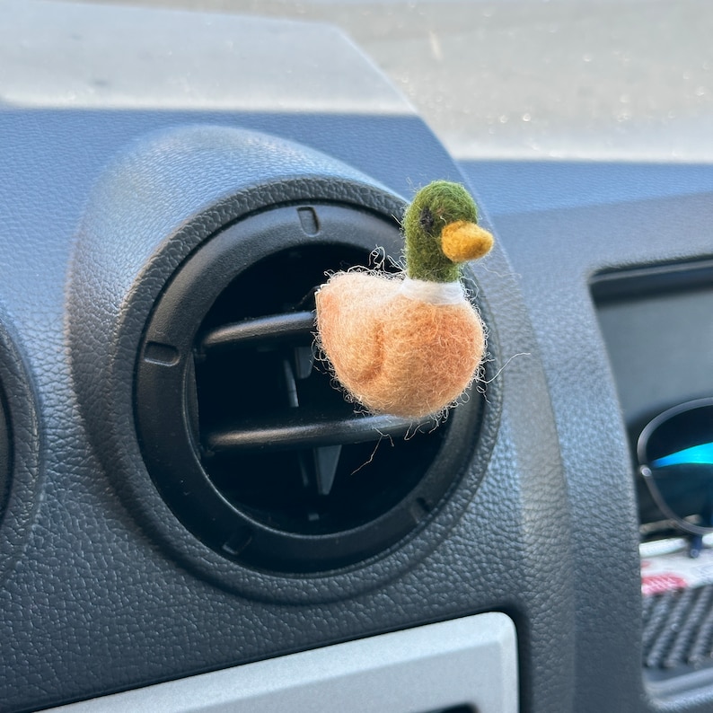 K&ouml;nnte beinhalten: Ein Filz-Enten-Lufterfrischer f&uuml;r das Auto mit gr&uuml;nem Kopf, braunem K&ouml;rper und gelbem Schnabel. Die Ente h&auml;ngt an einer L&uuml;ftung im Auto.
