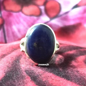 May include: A silver ring featuring a large, oval-shaped, deep blue lapis lazuli gemstone with subtle gold-colored flecks. The metal band has a split design on the sides. The text @samajit is printed in small white letters at the base of the stone.