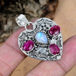 May include: A heart-shaped silver pendant featuring a central pear-cut iridescent moonstone surrounded by three oval-cut deep pink rubies. The silver surface is decorated with intricate stamped patterns and includes a bail at the top for hanging.