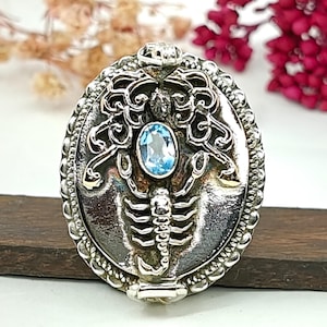 Può includere: Un ciondolo ovale in argento con un rilievo di scorpione e un bordo in filigrana intricato. Una singola pietra preziosa ovale sfaccettata di colore azzurro è incastonata al centro del corpo dello scorpione. Il ciondolo ha una finitura metallica.