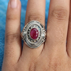 Può includere: Un anello in argento con una pietra preziosa rosso scuro di forma ovale e sfaccettata al centro. L'incastonatura è circondata da un bordo decorativo con una trama a corda intrecciata e piccoli accenti di perline in argento.