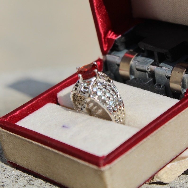 May include: A silver ring with a red gemstone set in a filigree design. The ring is in a red velvet ring box.