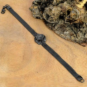 Puede incluir: Una pulsera de malla negra con una pieza central decorativa. La pulsera presenta un cierre de gancho y una pieza central decorativa con una piedra oscura. La pulsera se exhibe sobre una superficie de madera.
