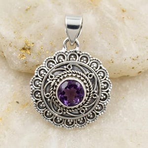 May include: A round silver pendant featuring a central faceted purple amethyst gemstone. The stone is surrounded by a twisted rope border and an outer scalloped frame with intricate filigree scrollwork and small silver bead accents.
