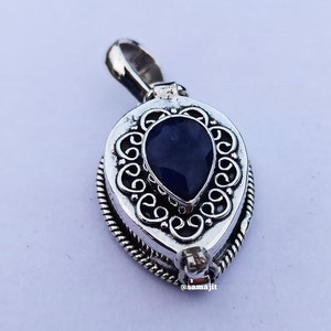 May include: A silver-colored pendant with a teardrop-shaped, dark blue gemstone at its center. The pendant has an ornate, swirling design around the gemstone and a textured border. The bail is at the top, and the pendant is on a white background.