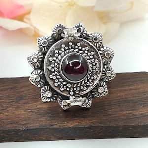 May include: A silver ring with a round, ornate design featuring a dark red gemstone in the center. The ring has a hinged opening and is decorated with small, raised details.