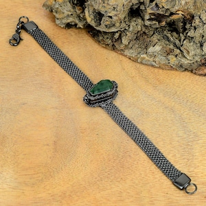 Puede incluir: Una pulsera de malla gris oscuro con una piedra preciosa verde rectangular en el centro. La pulsera tiene un cierre de gancho y un cierre de anillo. La piedra preciosa está engastada en un marco de metal gris oscuro.
